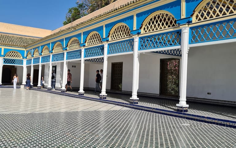 Sunlit courtyard at Bahia Palace in Marrakech, with blue-and-white arches, geometric tile flooring, and palm trees above the roofline.