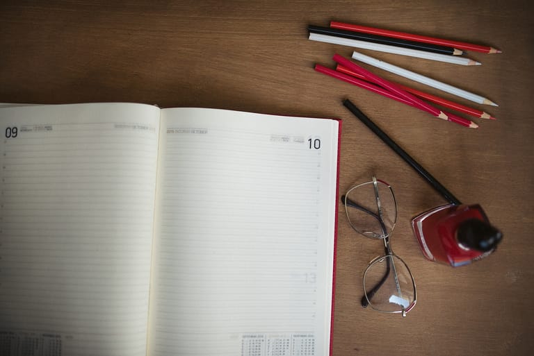 An open planner, pencils, glasses and pen arranged across a warm wooden desk
