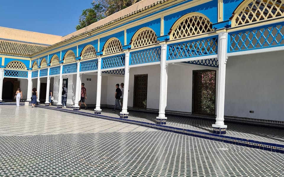 Sunlit courtyard at Bahia Palace in Marrakech, with blue-and-white arches, geometric tile flooring, and palm trees above the roofline.